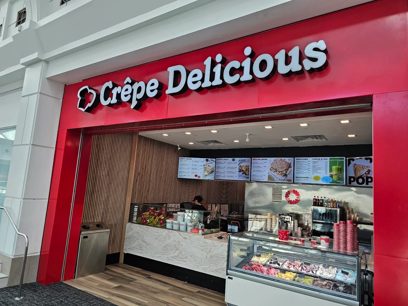 The finished Crepe Delicious unit at Menlo Park Mall — red illuminated signage fascia, slatted timber wall, menu boards, marble-front service counter and gelato display case.